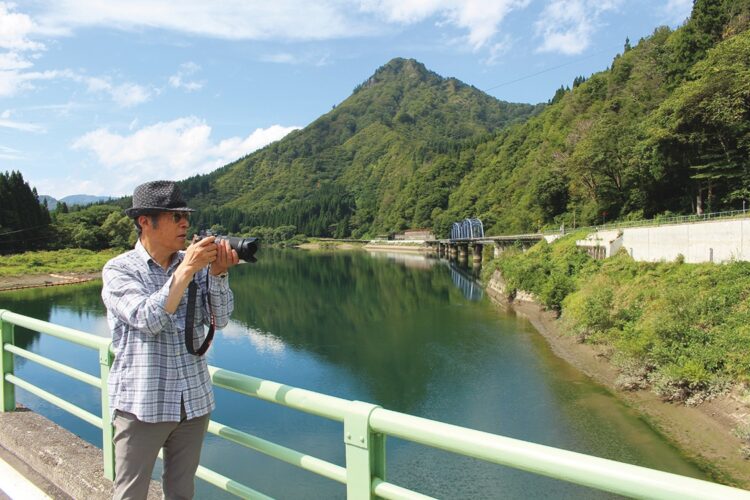 カメラを構える星さん。背景は11年ぶりに再開通した第8只見川橋梁